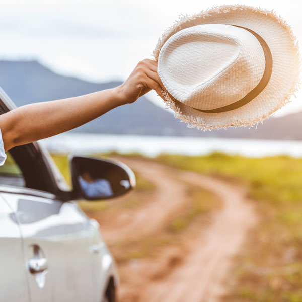 Happy woman hand holding hat outside open window car with meadow and mountain lake background. People lifestyle relaxing as traveler on road trip in holiday vacation. Transportation and travel concept